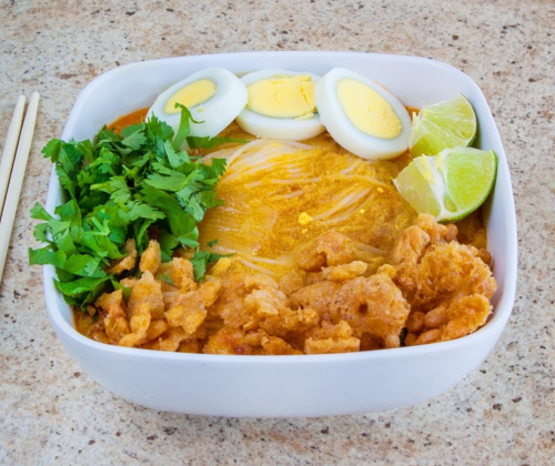 A bowl of noodles topped with fried chicken, boiled eggs, cilantro, and lime wedges on a textured surface.
