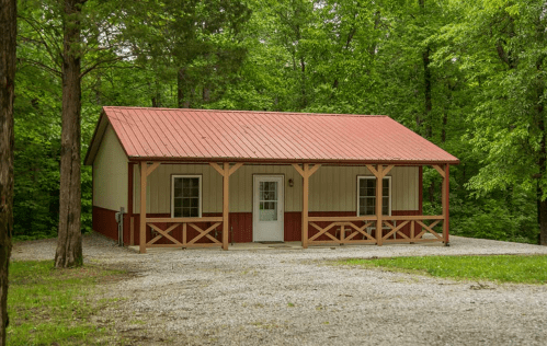 A small, rustic cabin with a red metal roof, surrounded by trees and gravel, featuring a porch and white door.