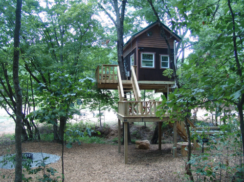 A wooden treehouse elevated among trees, featuring a deck and surrounded by a natural, wooded area.
