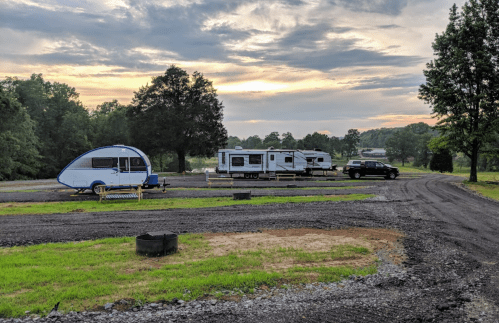 A scenic campsite with two trailers parked, surrounded by trees and a cloudy sky at sunset.