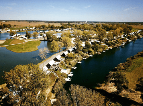 Aerial view of a serene marina with boats, surrounded by trees and open fields under a clear blue sky.