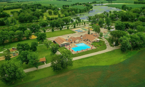 Aerial view of a resort with a pool, surrounded by green fields, trees, and a pond in a rural landscape.