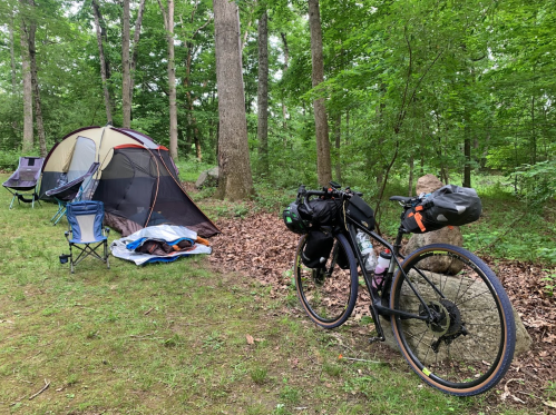 A campsite in a forest with a tent, camping chairs, and a bicycle loaded with gear.