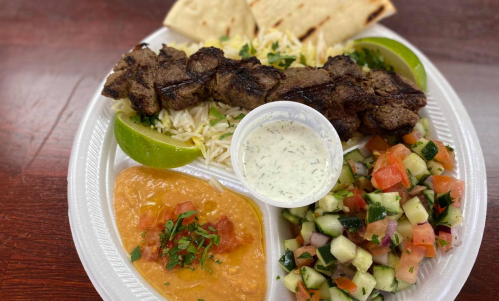 A plate of grilled meat skewers with rice, salad, lime, pita bread, and sauces, including a side of hummus.