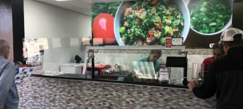 A busy restaurant counter with staff preparing food, customers waiting, and colorful salad images on the wall.