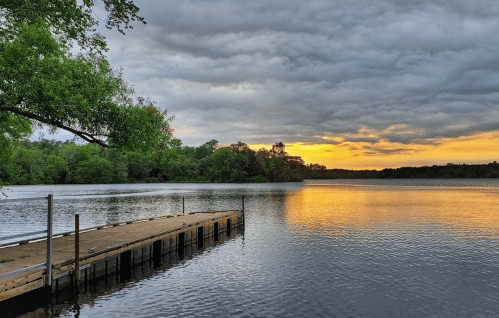 A serene lake at sunset, with a wooden dock extending into the water and dramatic clouds overhead.
