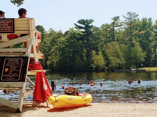 Lifeguard tower by a lake with swimmers enjoying the water on a sunny day, surrounded by trees.