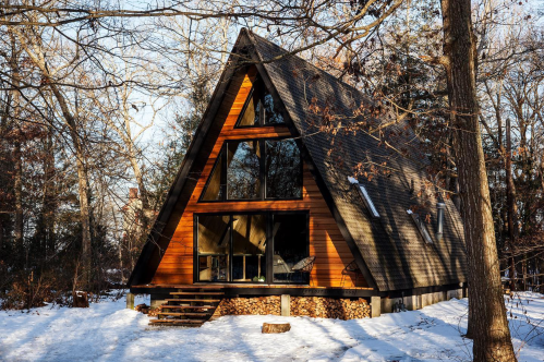A cozy A-frame cabin in a snowy forest, featuring large windows and stacked firewood beneath.