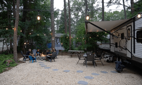 A cozy outdoor gathering with friends around a fire pit, surrounded by trees and an RV, with string lights overhead.