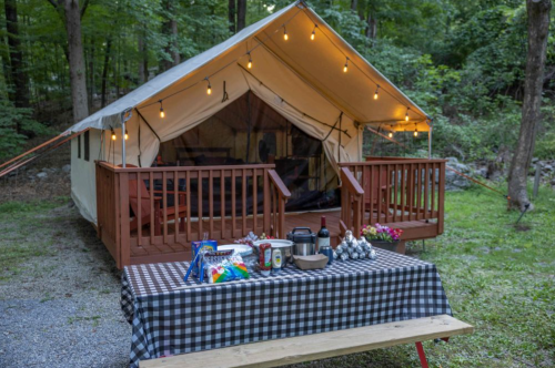 A cozy glamping tent with string lights, surrounded by trees, and a picnic table set with snacks and drinks.