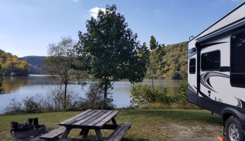 A serene lakeside view with a picnic table, tree, and an RV parked nearby, surrounded by autumn foliage.