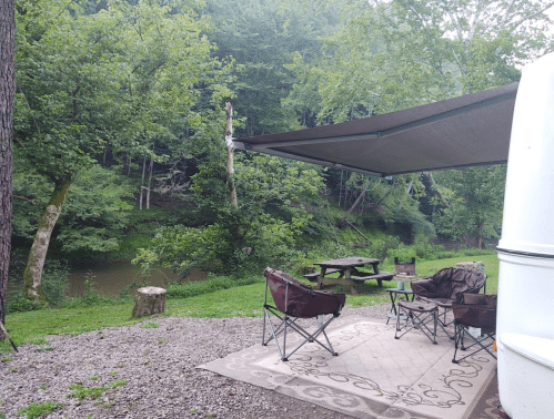 A peaceful campsite by a river, featuring two chairs, a picnic table, and a shaded area under a canopy.