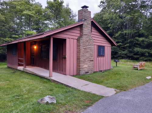 A rustic cabin with a stone chimney, surrounded by greenery and a gravel path leading to the entrance.