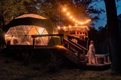 A cozy geodesic dome lit up at night, with a person standing on the steps and string lights illuminating the scene.