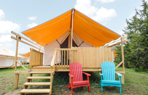 A glamping tent with an orange canopy, wooden deck, and two colorful chairs in front on a grassy area.