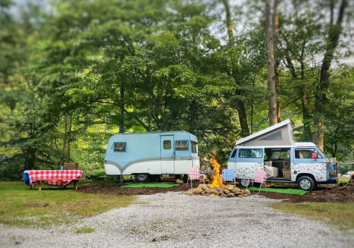 A cozy campsite featuring a vintage camper, a pop-up van, a picnic table, and a campfire surrounded by trees.