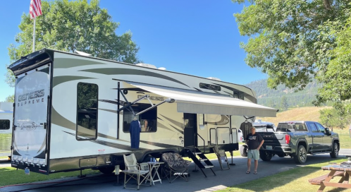 A large RV parked under trees, with an awning extended, and a truck nearby. A person stands outside, enjoying the scenery.