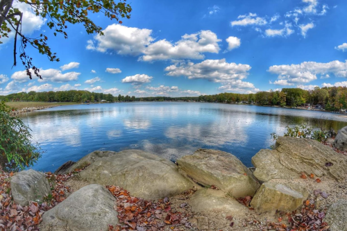 A serene lake scene with rocky shorelines, reflecting clouds and trees under a bright blue sky.