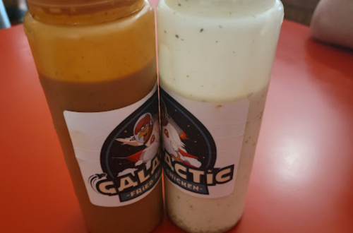 Two bottles of sauce, one brown and one white, with a "Galactic Fried Chicken" label, placed on a red table.