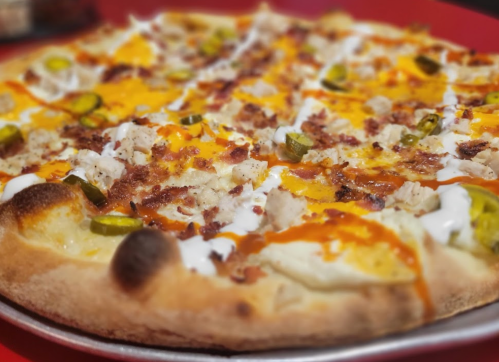 A close-up of a pizza topped with chicken, jalapeños, cheese, and drizzled with ranch and hot sauce on a red table.