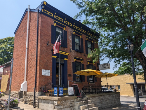 A brick building with yellow trim, featuring an American flag and outdoor seating, houses Strong's Pizzeria.