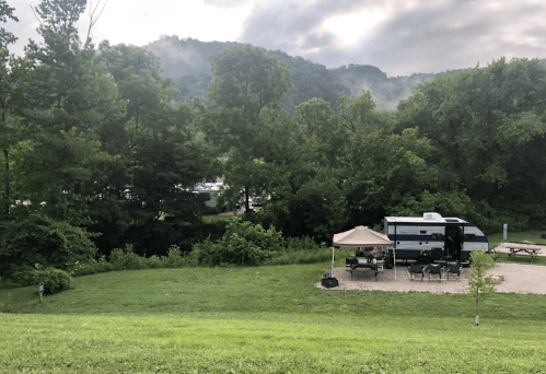 A scenic campsite with an RV, picnic area, and trees, set against a misty mountain backdrop.