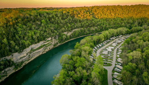 Aerial view of a river winding through lush greenery, with a campground of RVs along the water's edge.