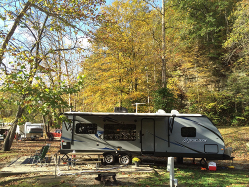 A gray travel trailer parked in a campsite surrounded by autumn trees and a rocky hillside.