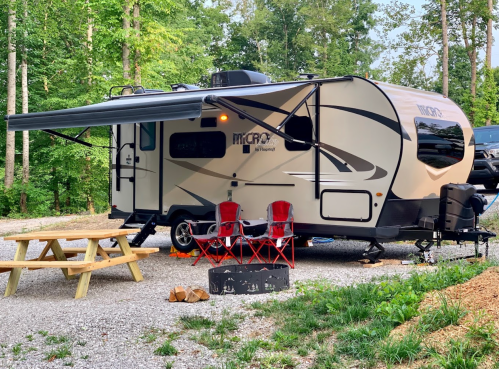 A small camper trailer parked in a wooded area, with a picnic table, chairs, and a fire pit nearby.