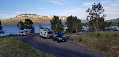 A scenic lakeside campsite with RVs and trees, surrounded by mountains under a clear blue sky.
