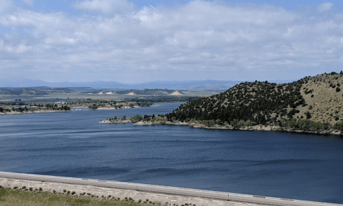 A serene lake surrounded by green hills and distant mountains under a partly cloudy sky.