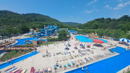 A vibrant water park scene with slides, pools, and sunbathers enjoying a sunny day amidst green hills.