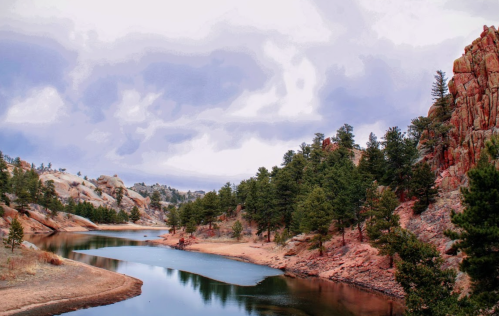 A serene river winds through rocky terrain, surrounded by pine trees under a cloudy sky.