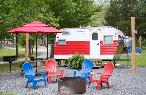 A vintage red and white camper surrounded by colorful chairs and a fire pit under a red umbrella in a wooded area.