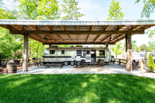 A vintage Airstream trailer parked under a wooden shelter, surrounded by green grass and trees.