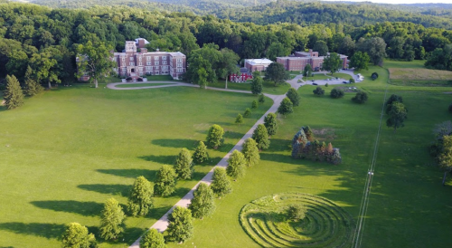 Aerial view of a green landscape featuring a large building, tree-lined paths, and a circular maze in the grass.