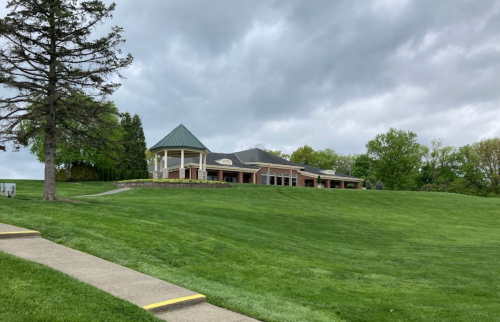 A grassy hill with a modern building in the background under a cloudy sky. A tree stands to the left.