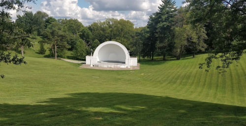 A white band shell sits on a grassy hill surrounded by trees under a partly cloudy sky.