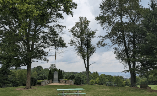 A scenic park view with two trees framing a stone monument and a picnic table, overlooking a distant landscape.
