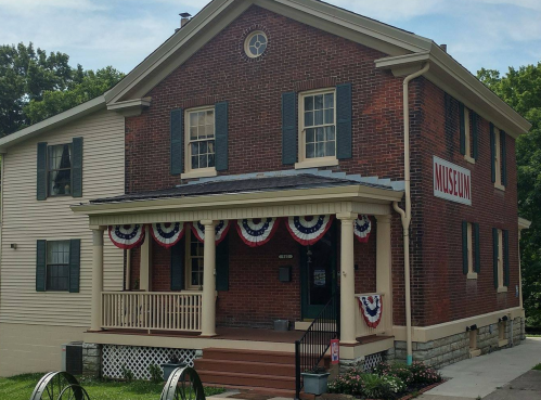 A brick building with blue shutters and patriotic decorations, labeled "MUSEUM," surrounded by greenery.