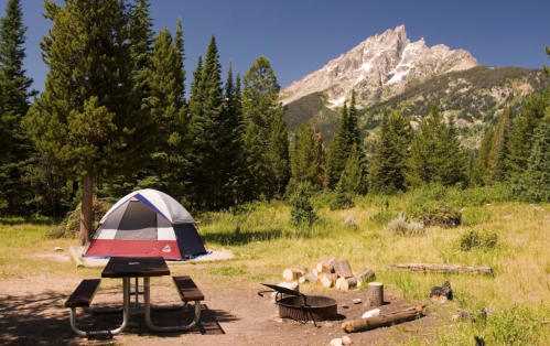 A colorful tent set up in a forest clearing, with a picnic table and fire pit, surrounded by mountains and trees.