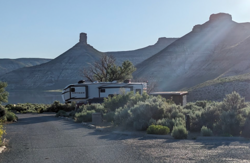 A camper parked near a winding road, surrounded by sagebrush and mountains under a clear blue sky.