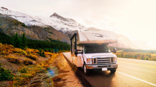 A white RV parked on a scenic road with mountains and trees in the background, illuminated by soft sunlight.