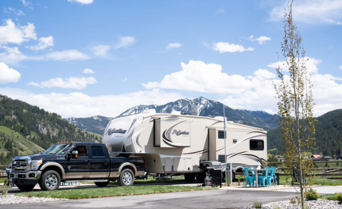A truck parked next to a large RV in a scenic mountain landscape with blue skies and green grass.