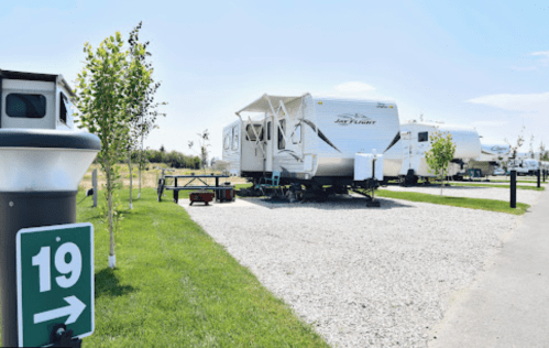 A parked RV in a grassy area with a gravel driveway, surrounded by trees and other RVs in the background.