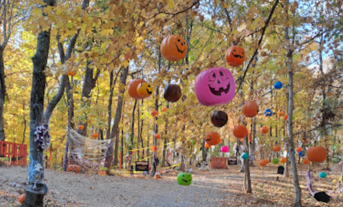 Colorful Halloween decorations, including hanging pumpkins, adorn a wooded path surrounded by autumn foliage.