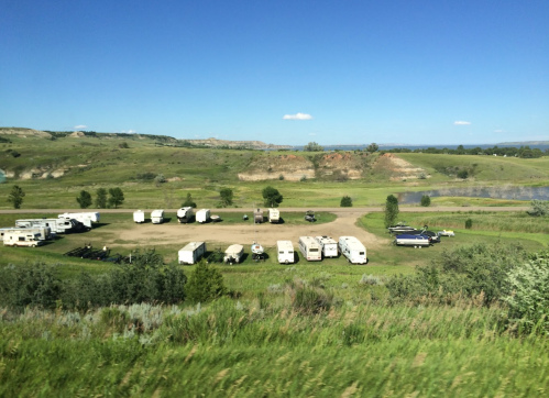 A scenic view of a grassy area with several RVs parked, surrounded by hills and a clear blue sky.