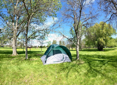 A green and gray tent set up on lush green grass, surrounded by trees under a clear blue sky.