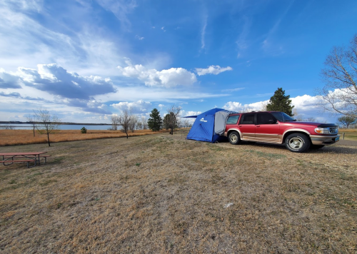 A red SUV parked near a blue tent by a lake, with grassy land and scattered trees under a cloudy sky.
