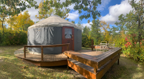 A cozy yurt on a wooden deck surrounded by trees and colorful autumn foliage under a partly cloudy sky.